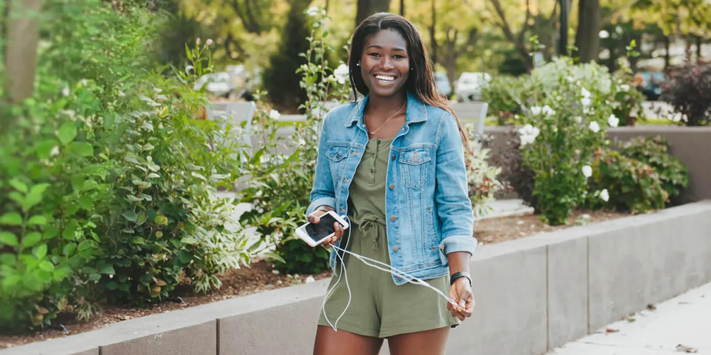 Young black woman wearing jean jacket and holding phone and headphones outside