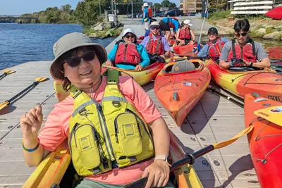 A woman in sunglasses and a hat smiles for a photo while sitting in a kayak on a dock. Several other people are behind her in kayaks.