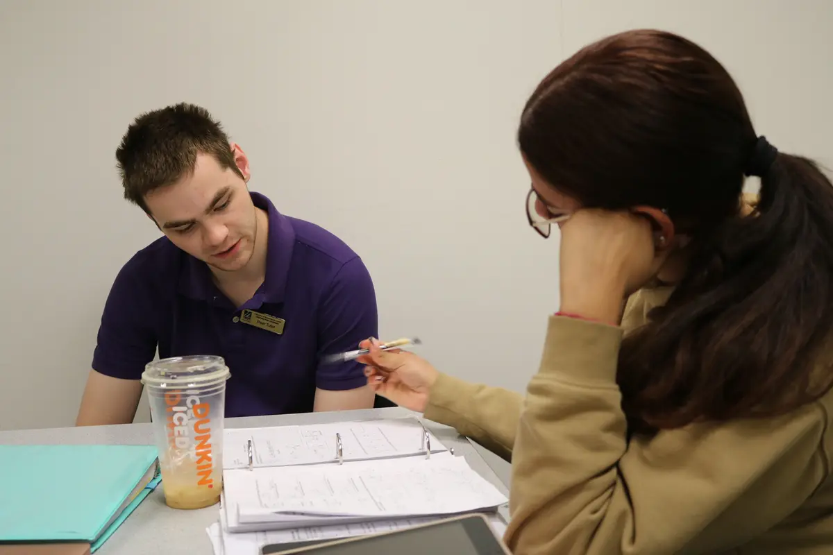 A tutor and student, seated, look over a notebook and discuss class work.