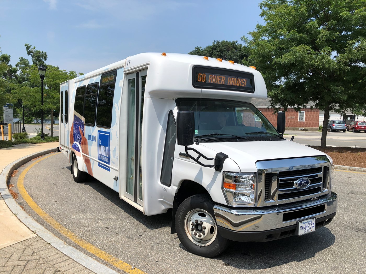 A white UMass Lowell shuttle stopped at a bus stop on South Campus. The LED sign in the front reads "Go Riverhawks".