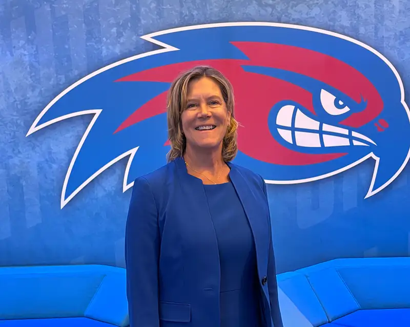 Amy Hoey standing in front of a UMass Lowell River Hawks (athletics) logo.