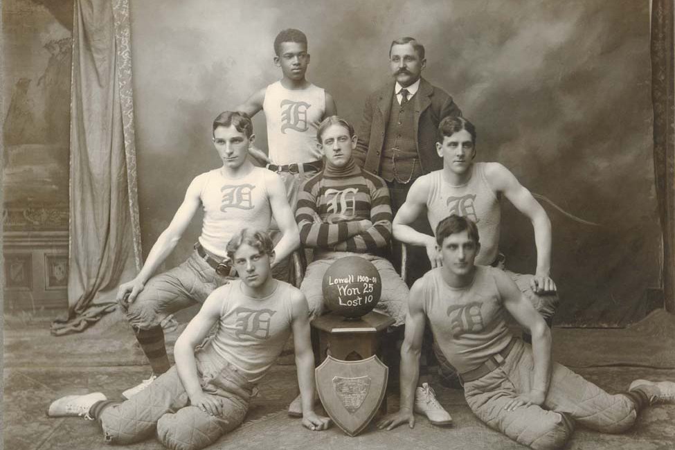 A basketball team of six young men and a coach pose for a photo with a basketball dated 1900-01.