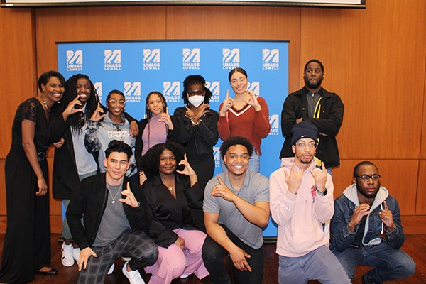 A dozen young men and women pose for a group photo in front of a blue UMass Lowell backdrop