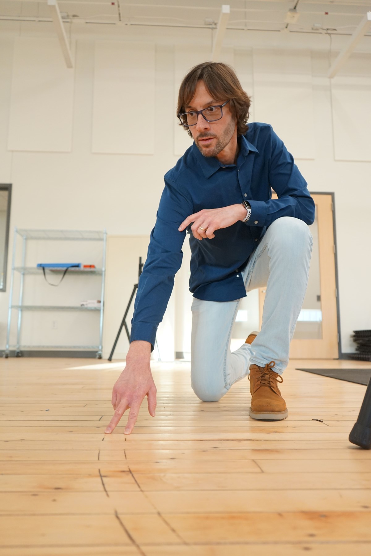 A man wearing glasses kneels on a hardwood floor and touches faint lines with his fingers