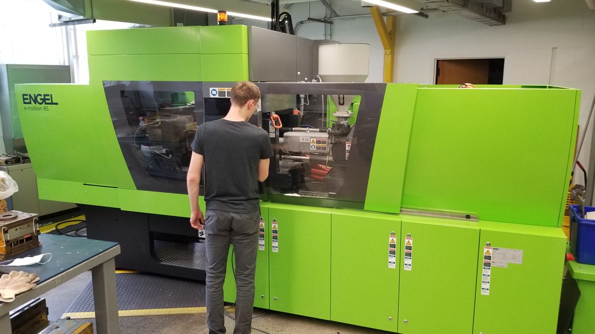 Male student looking into Engel e-motion 85 injection molding machine. It is described as: The ENGEL e-motion is an all-electric injection moulding machine for high-end applications. The right choice when both precision and speed are essential.