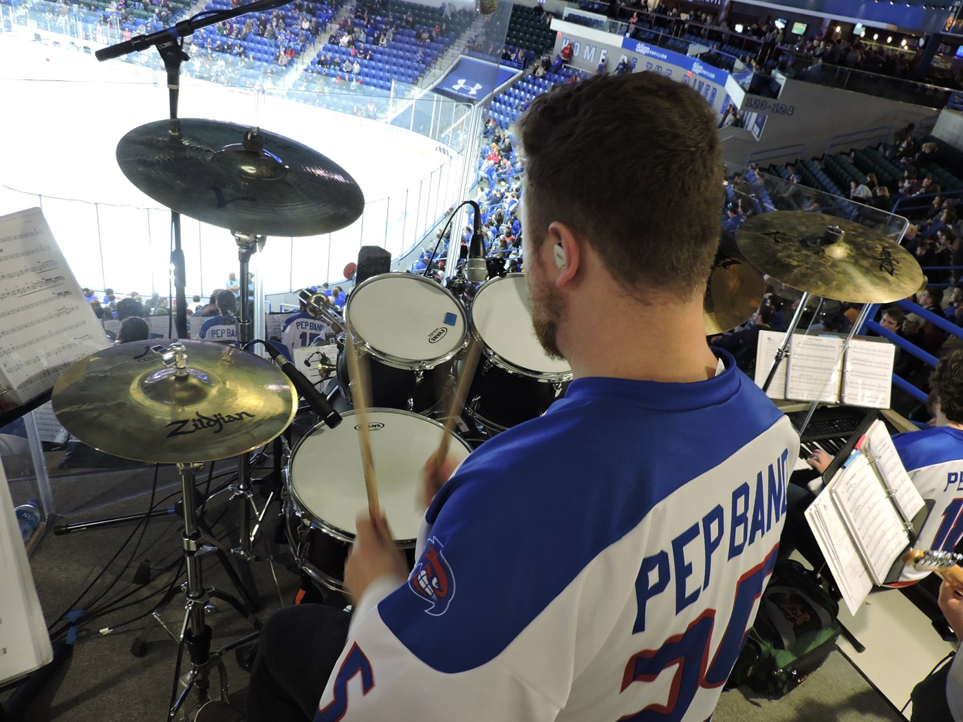 Drumset musician in hockey pep band shirt overlooks the entire pep band and ice surface at the Tsongas Center on UMass Lowell's campus where the River Hawks ice hockey team is competing.