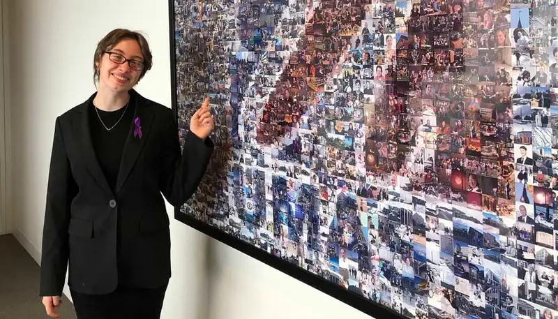 Meghan Dearing stands in front of a mosaic of small photos that are arranged to form the UMass logo.