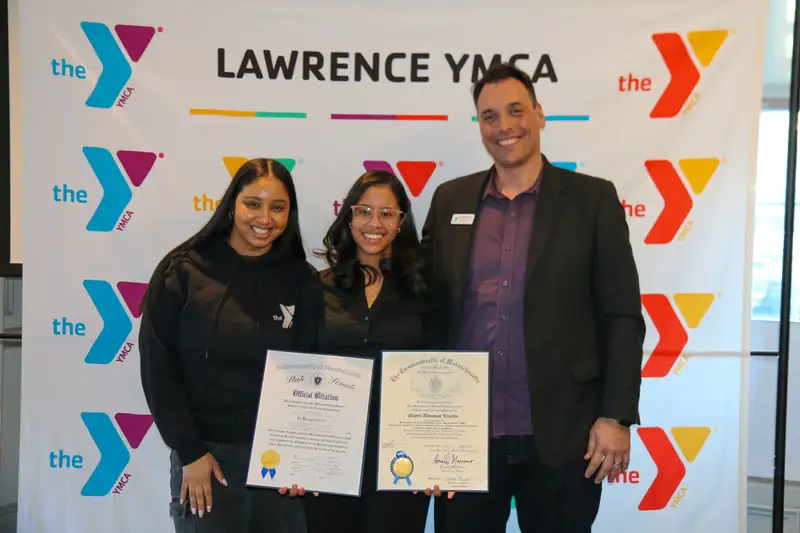 Nayeli Almanzar Lizardo stands with two other people while holding certificates in front of a Lawrence YMCA banner.