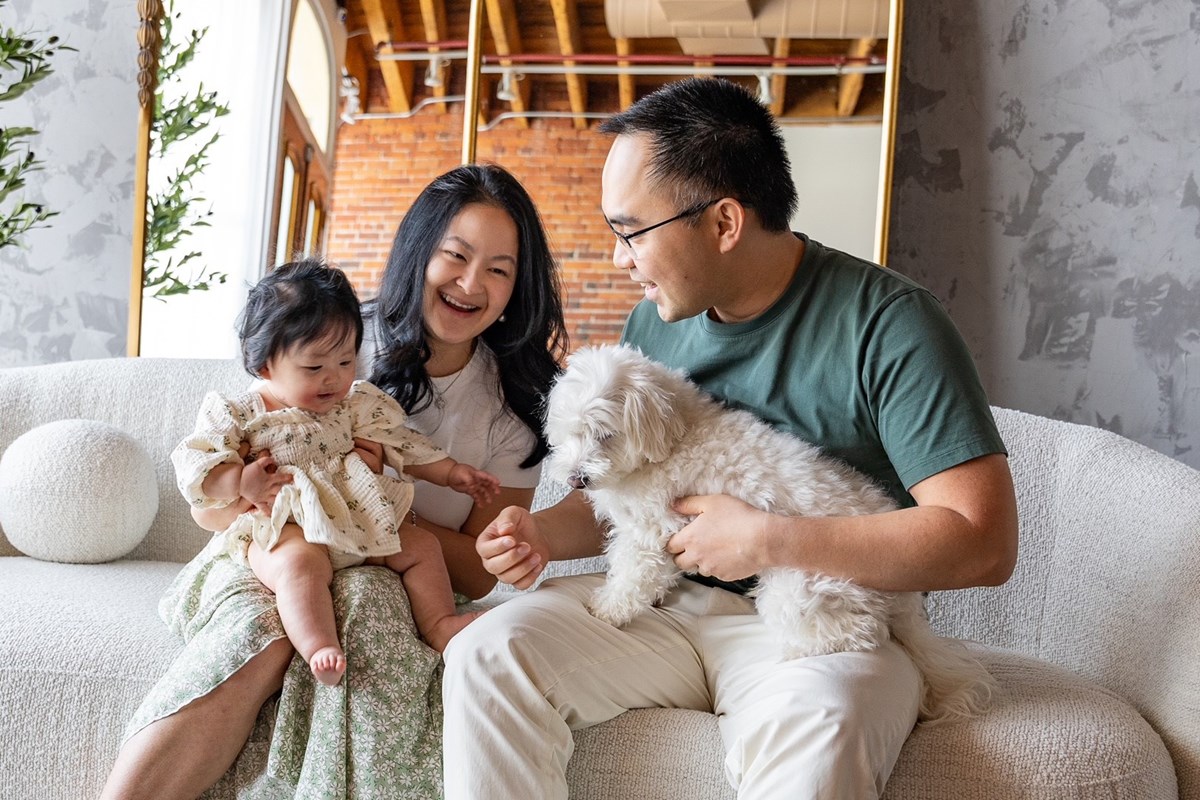 A husband and wife hold their young daughter and a dog while sitting on a couch.