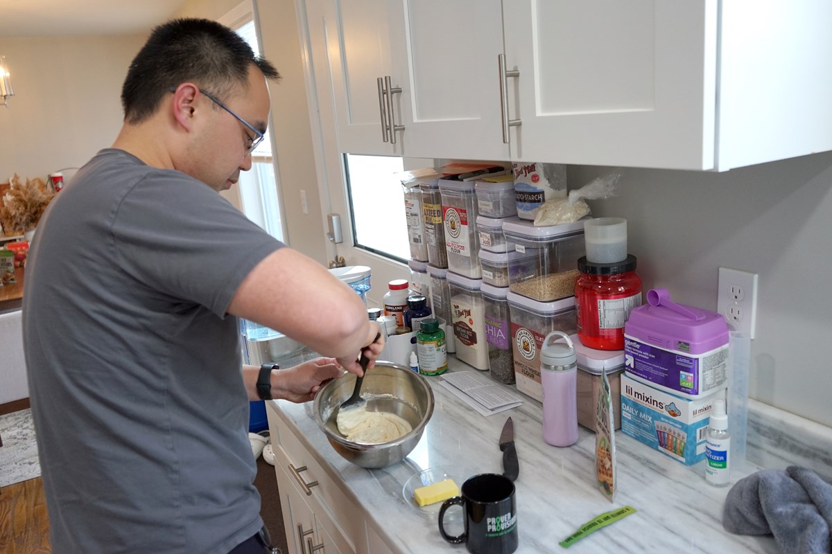 A man in a gray T-shirt mixes waffle batter in a silver bowl in a kitchen.