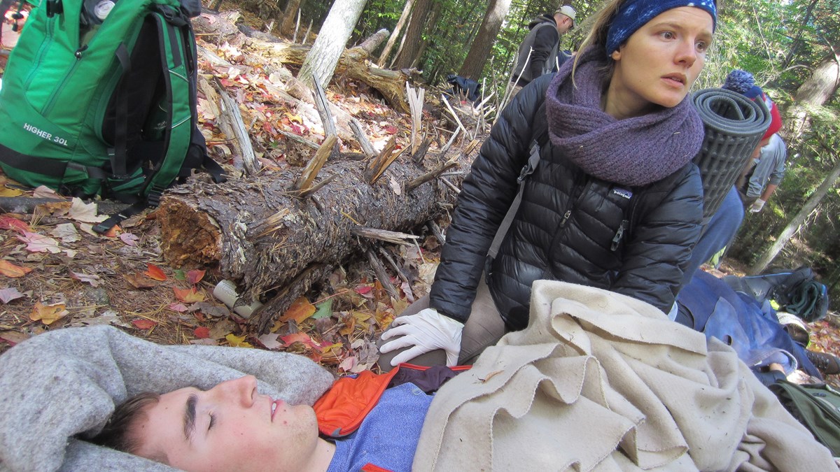 Picture of a Wilderness First Aid class emergency scenario with woman wearing gloves looking up and helping man on ground wrapped in blankets