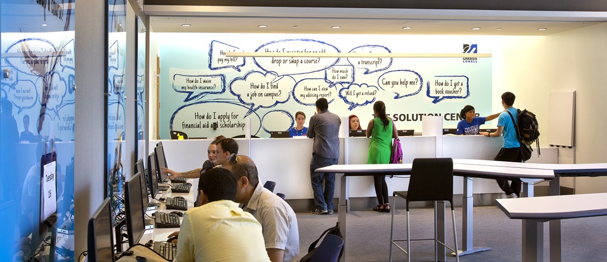 Exterior of Umass Lowell Solution Center, group of students getting assistance at desk and computers from Solution Center members.