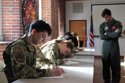 A group of UMass Lowell AFROTC cadets sits in a line at desks writing on paper with pencils as an instructor looks on.