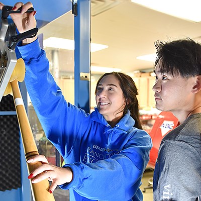 One mechanical engineering student reaches high up on a baseball bat testing machine while another student watches.