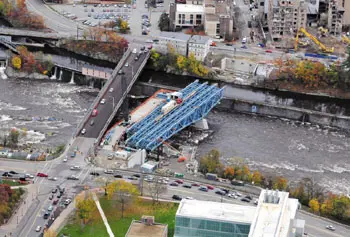 Blue steel girders for the new University Avenue bridge extend across the Merrimack River, soon to replace the 116-year-old Textile Memorial Bridge, to its left. At top, cranes are demolishing the former St. Joseph's Hospital, clearing way for UMass Lowell's new student center, seen as a hub uniting the north, east and south campuses.