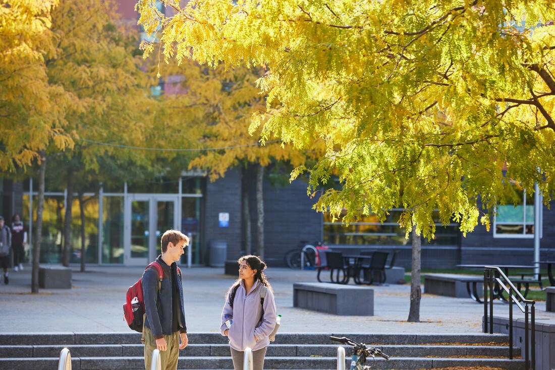 Male and female student wearing backpacks talk in front of academic building wit yellow trees behind