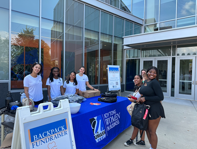 A group of female students stand in front of a table holding backpacks.