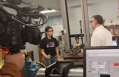 UMass Lowell’s Patrick Drane, assistant director of the Baseball Research Center, demonstrates the strength of a hockey stick with “NESN Clubhouse” show host, Bella. Credit: UMass Lowell