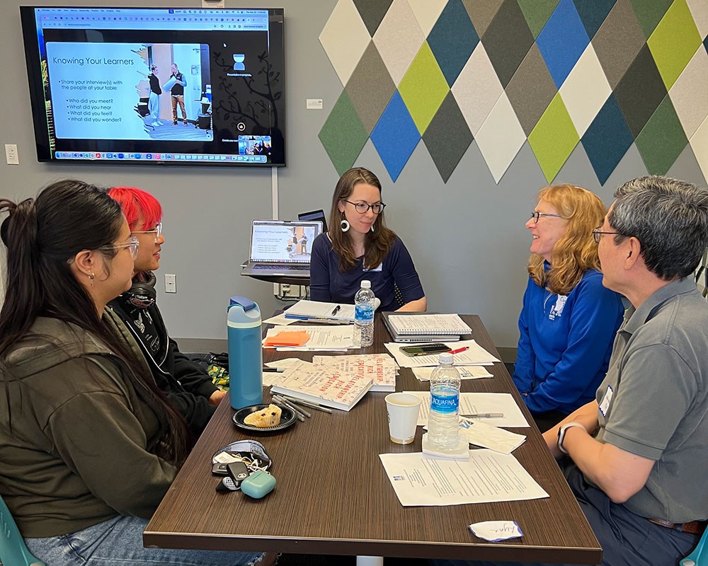 Instructors, sitting around a table, engage in a Leading, Engaging and Advising Fellows workshop.