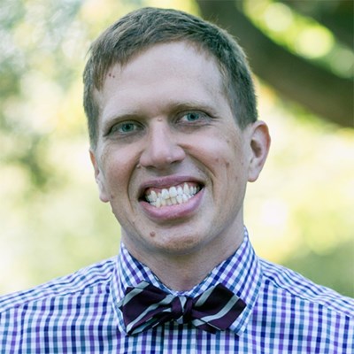 Caucasian man with blonde hair in a plaid shirt and bowtie smiles