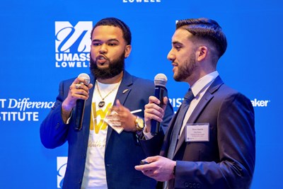 Two young men in blazers hold microphones and speak in front of a blue backdrop.