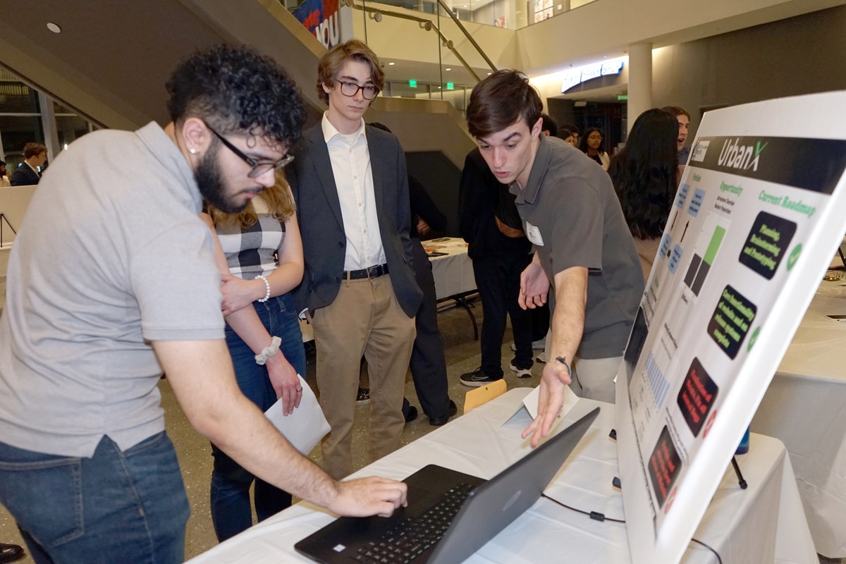 A young man gestures toward a laptop while another young man types on it as another young man looks on in a building lobby.