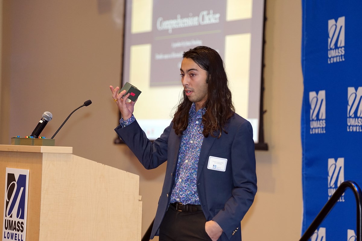 A student with long hair in a suit holds up a small black box while standing at a podium on a stage.