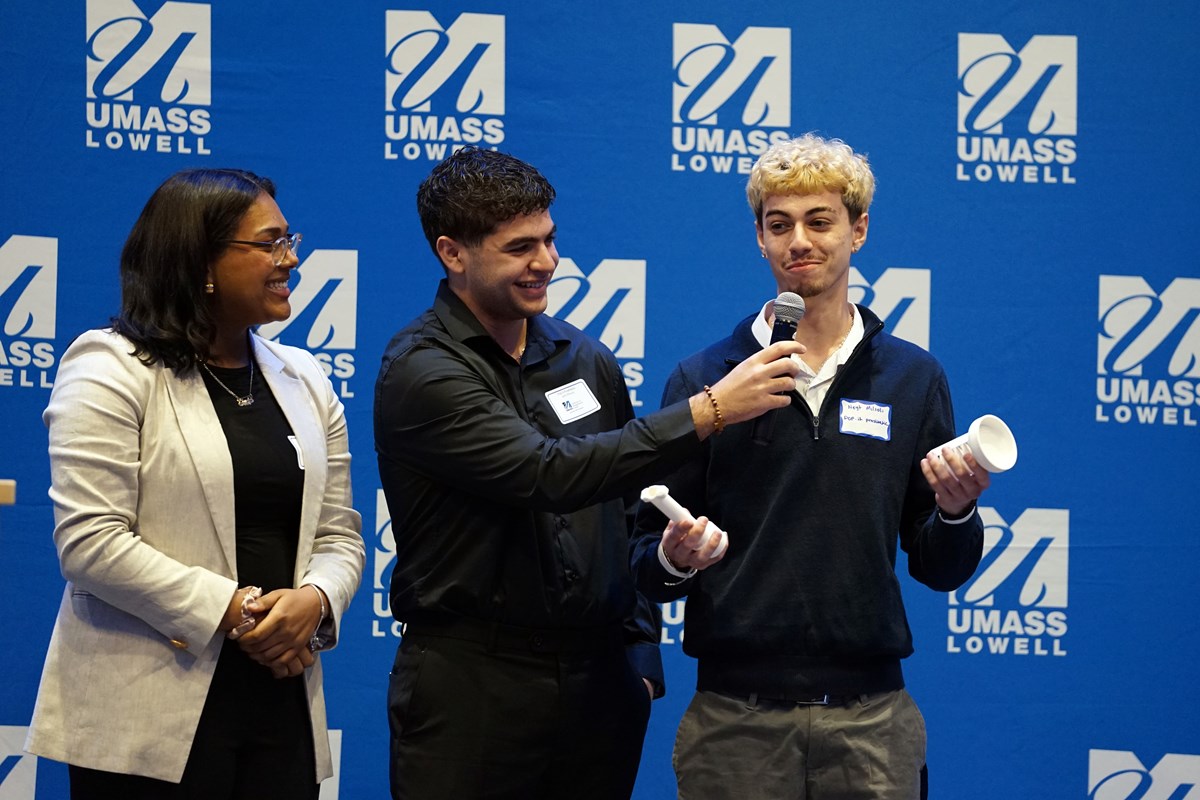 A student smiles while holding two pieces of plastic while another student holds a microphone in front of him and a third student looks on.