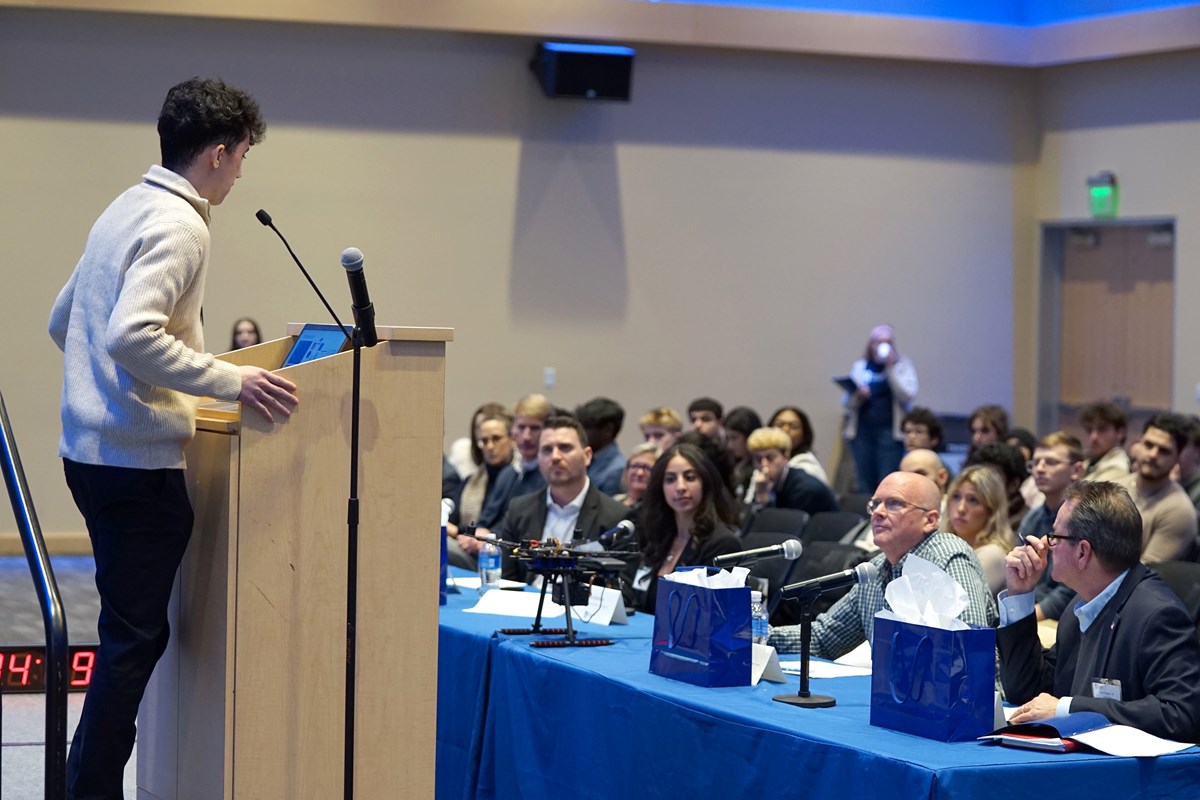 A young man stands behind a podium on stage and speaks to a crowd of people seated in a ballroom.