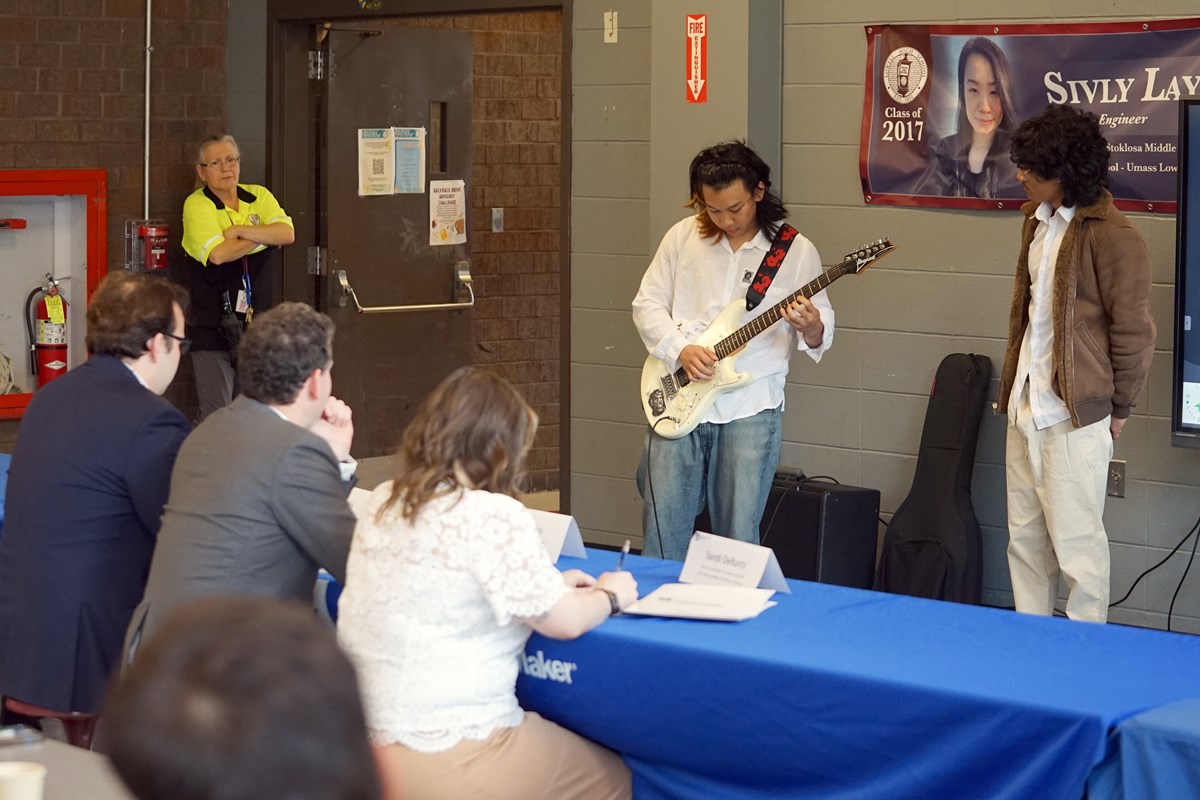 A person plays a guitar while several people look on in a school cafeteria.