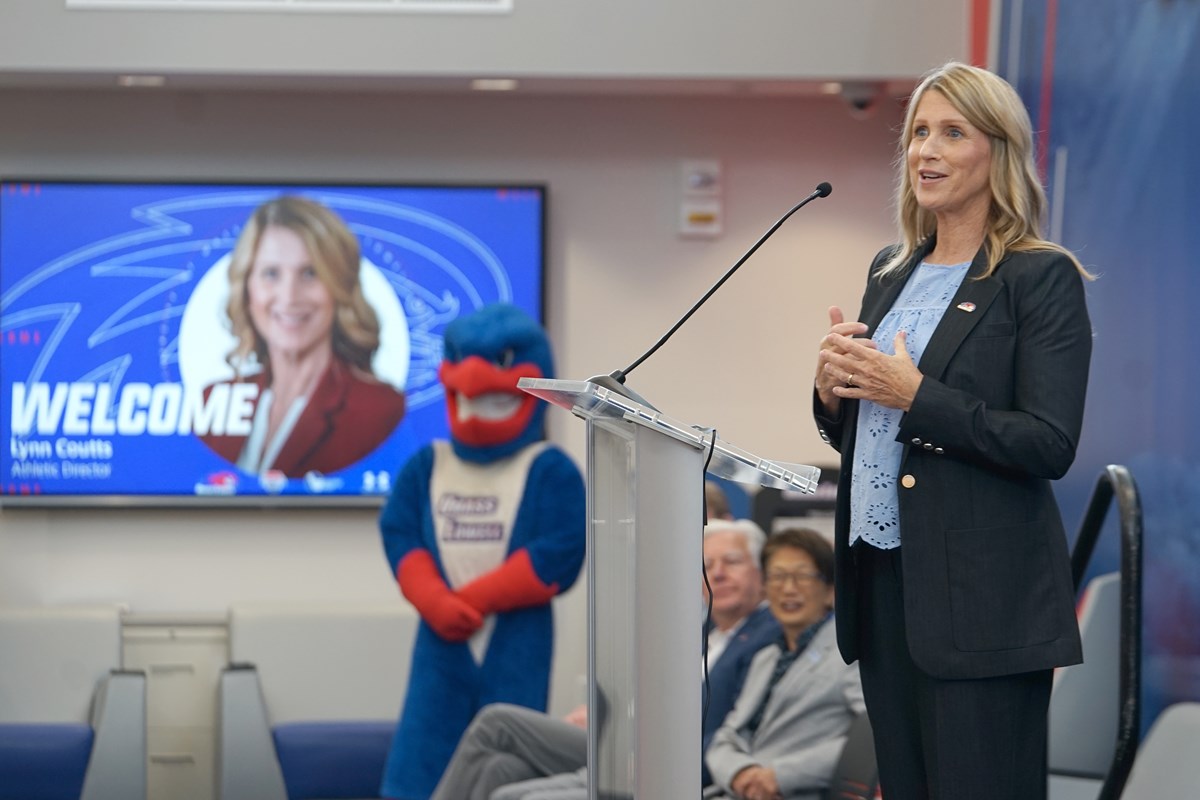 A woman speaks at a podium on a stage. In the distance are a college mascot and a screen with her visage.