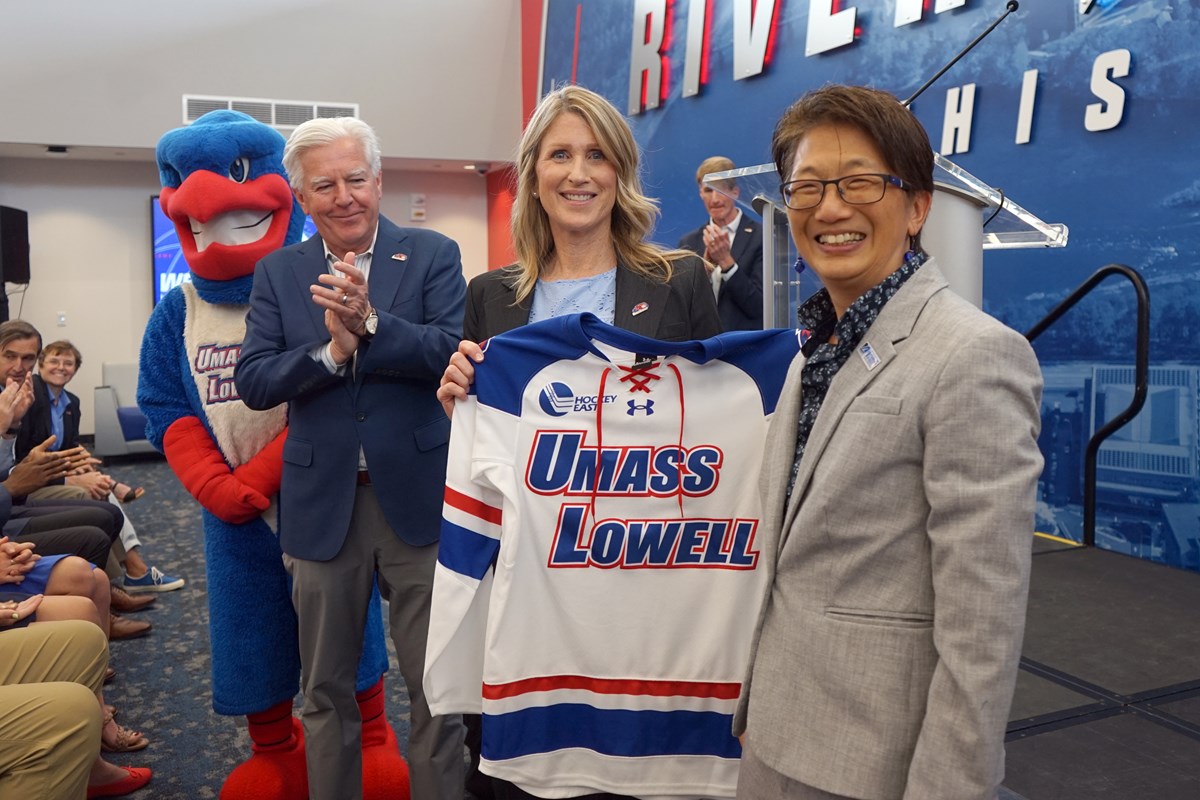 A woman holds a UML hockey jersey while standing between a man and a woman. A River Hawk mascot looks on.