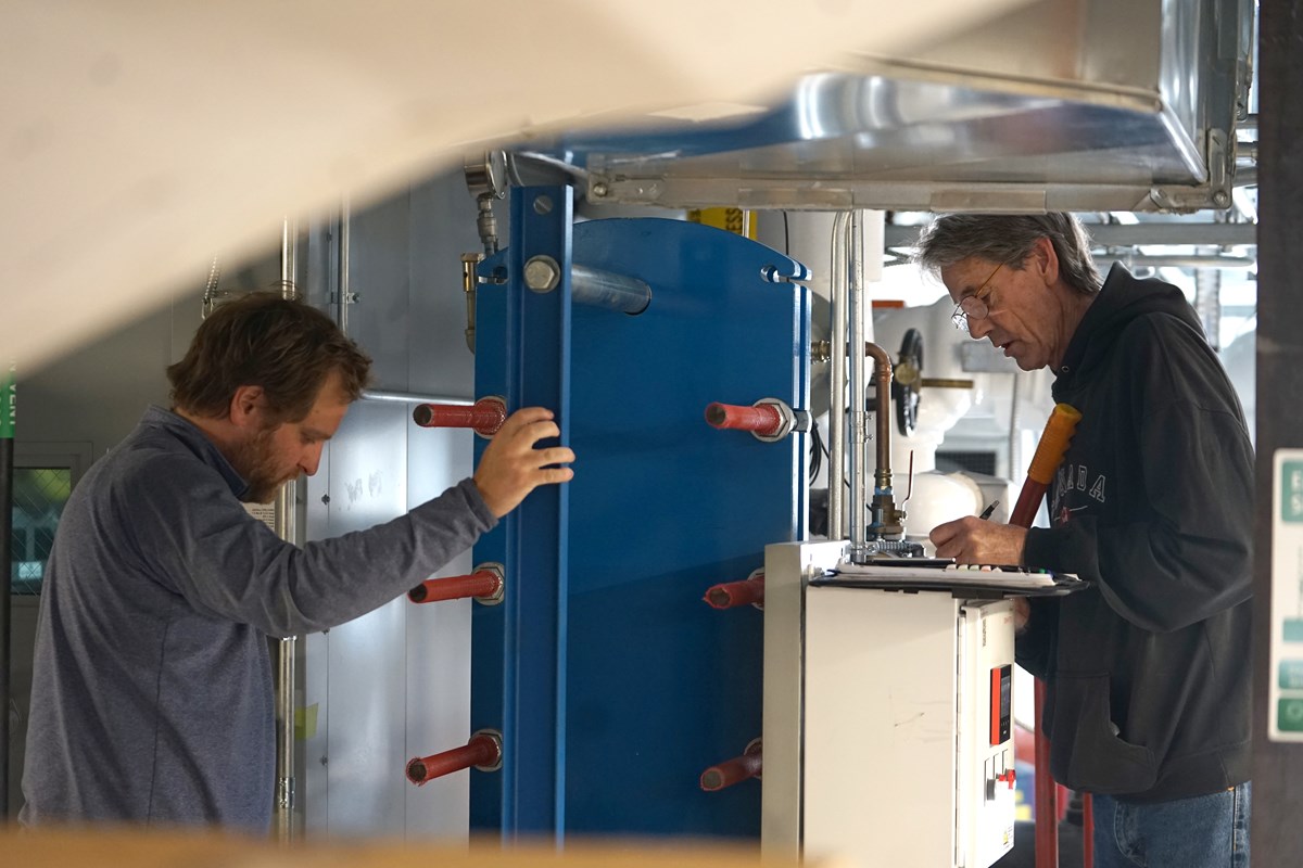 Two people look at a buiding system's equipment. One of them is writing.