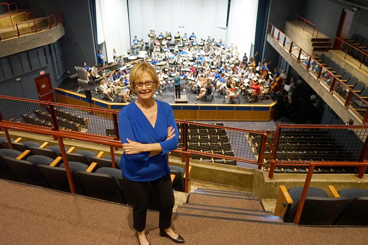 A woman stands on a theater balcony and poses with her arms folded. A band is on the stage below