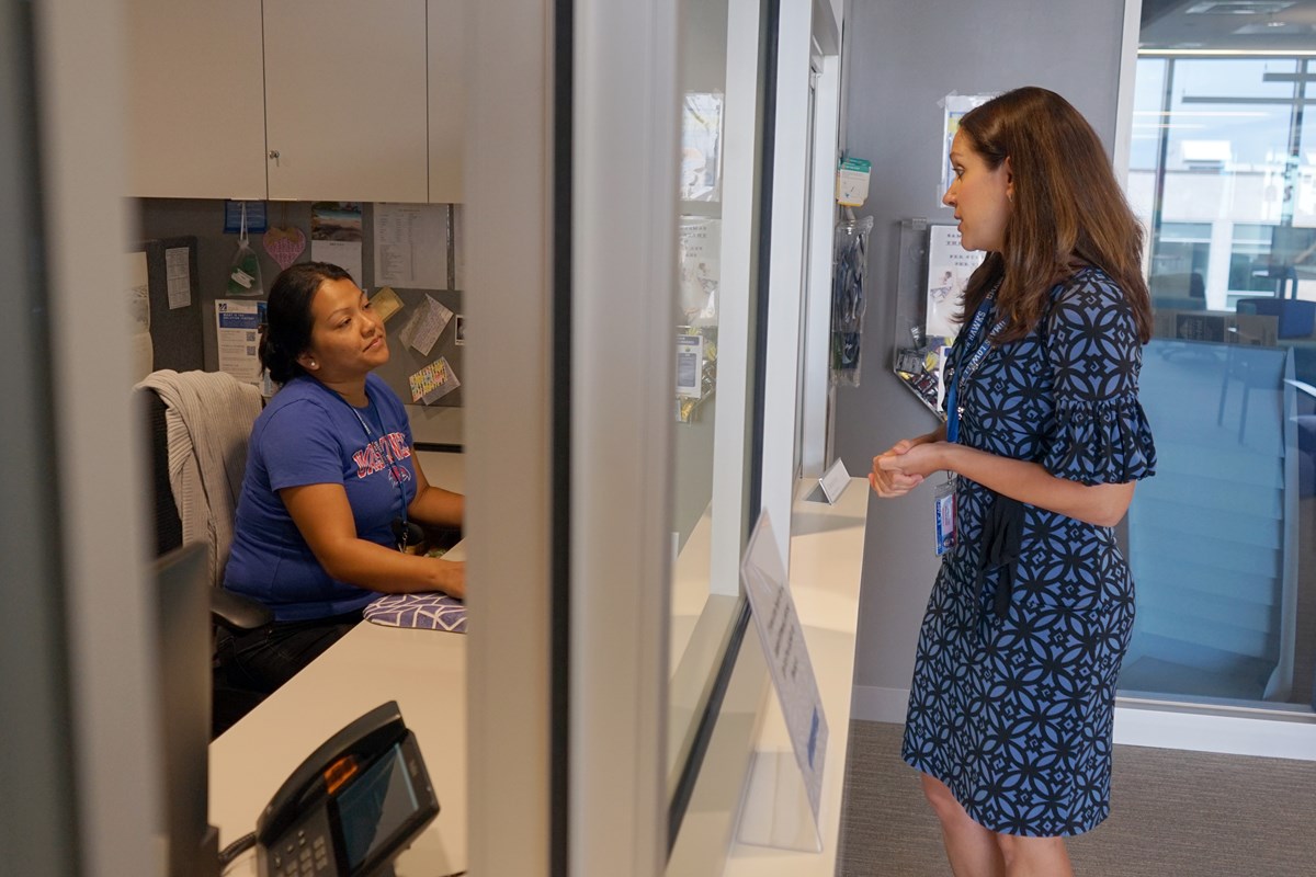 A person in a blue dress talks to a person in a blue T-shirt who is sitting at a desk behind a divider in an office.