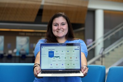 A student smiles while holding a laptop toward the camera.