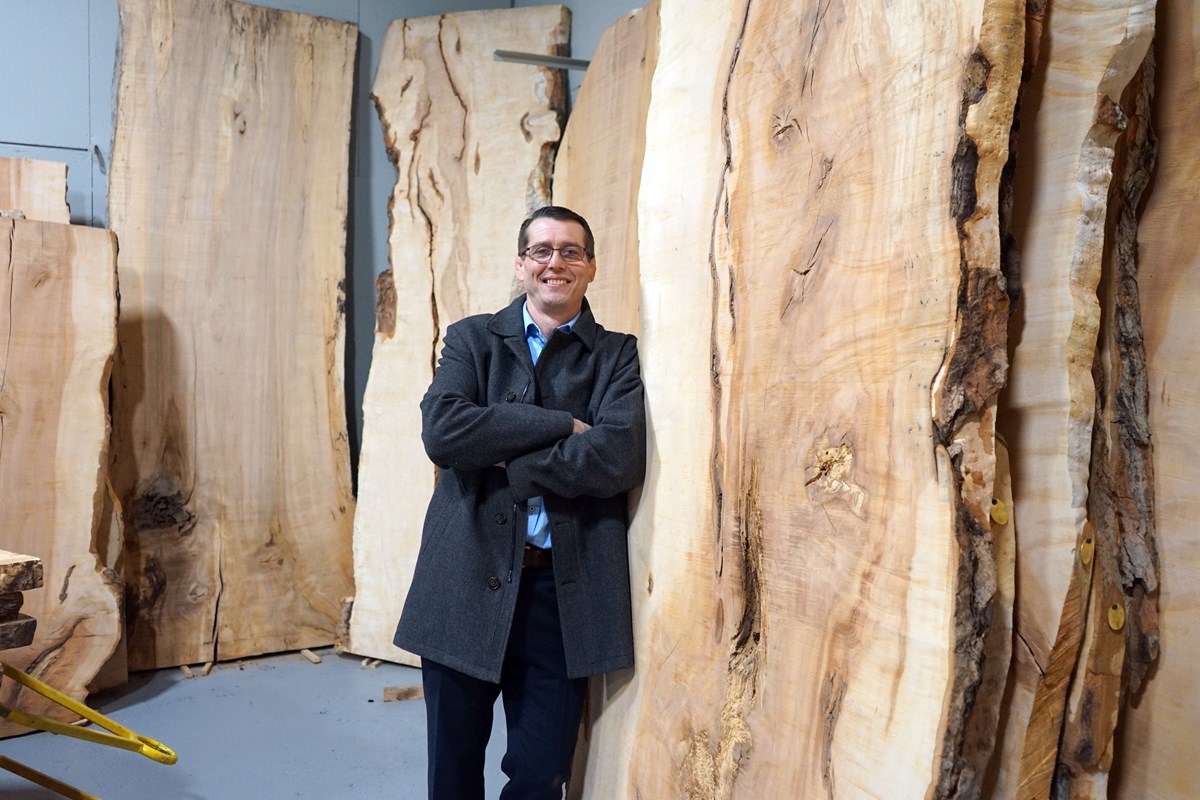 A person with glasses and a wool coat leans against slabs of wood in a wood shop.