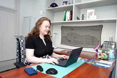 A woman with long hair and in a black top works at a laptop at her desk in her office.