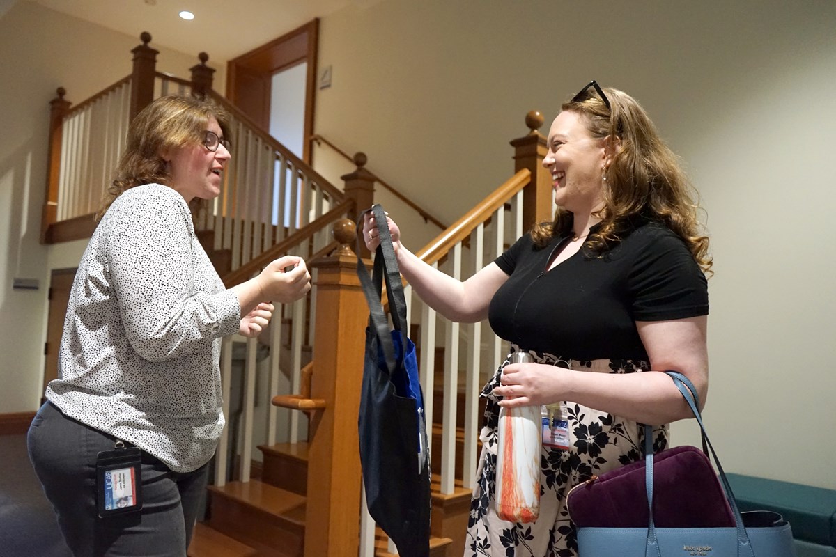 A woman smiles as she takes a bag from another woman while standing in front of a stairway bannister.