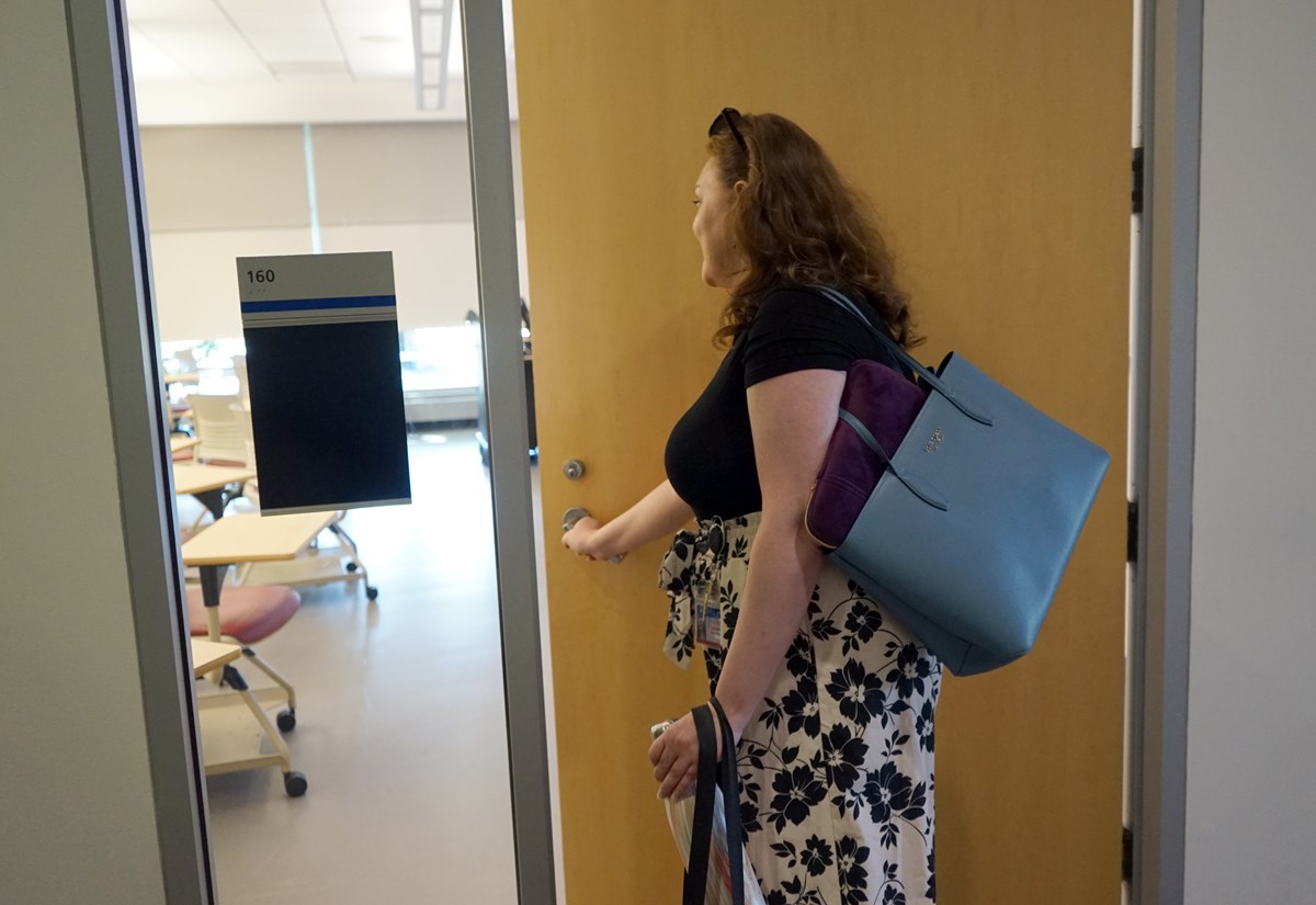 A woman with long hair and a blue purse over her shoulder opens a classroom door.