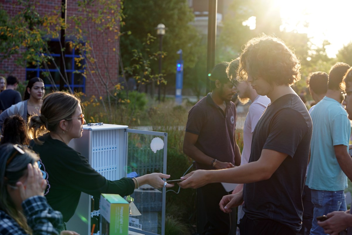 A young man hands his phone to a young woman at a table at an outdoor event at sunset.