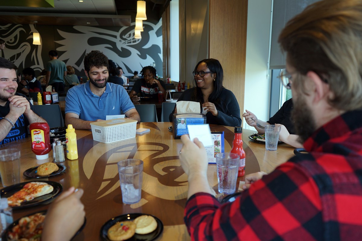 A group of people read from cards while sitting at a round table in a college dining hall.