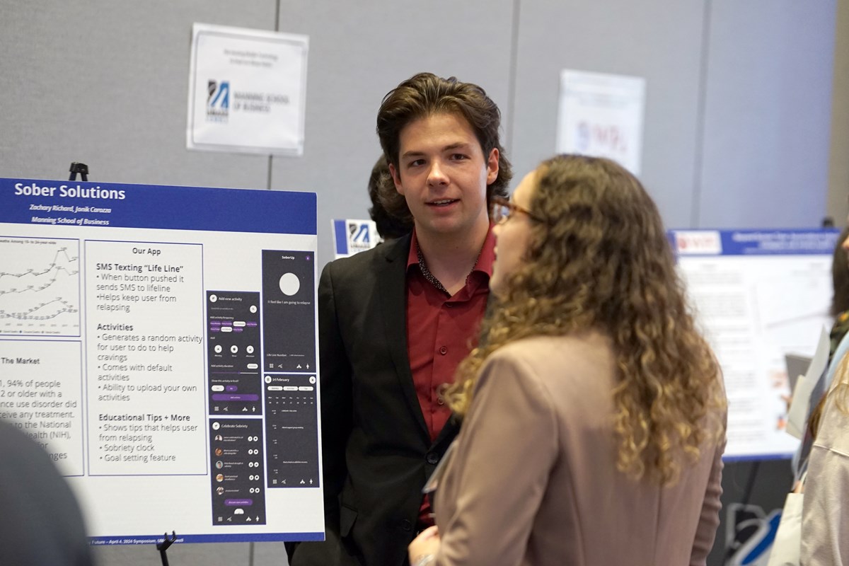 A young man with dark hair wearing a suit talks to a young woman who is looking at his poster at a symposium.