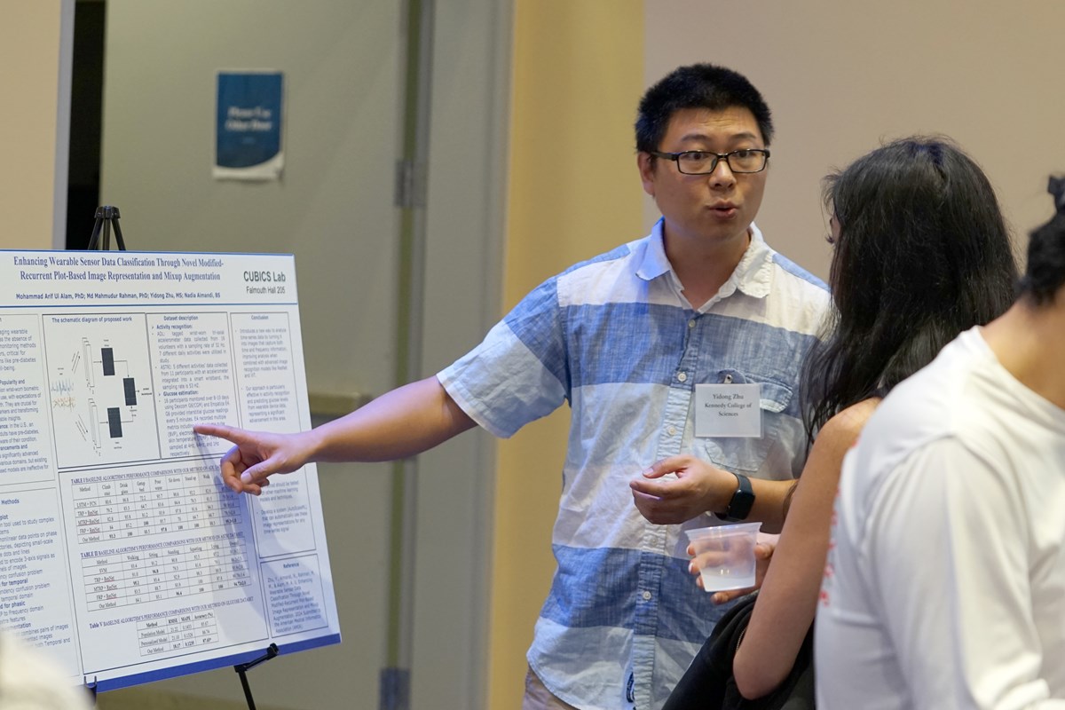 A man with dark hair and glasses points to a poster while talking to a woman at a symposium.