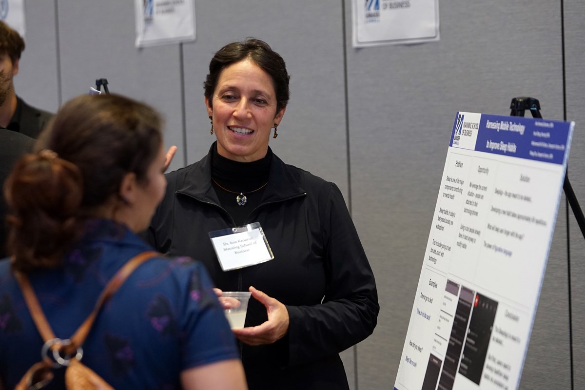 A woman with short hair and a dark outfit is holding a cup and talking to a person who is looking at her poster.