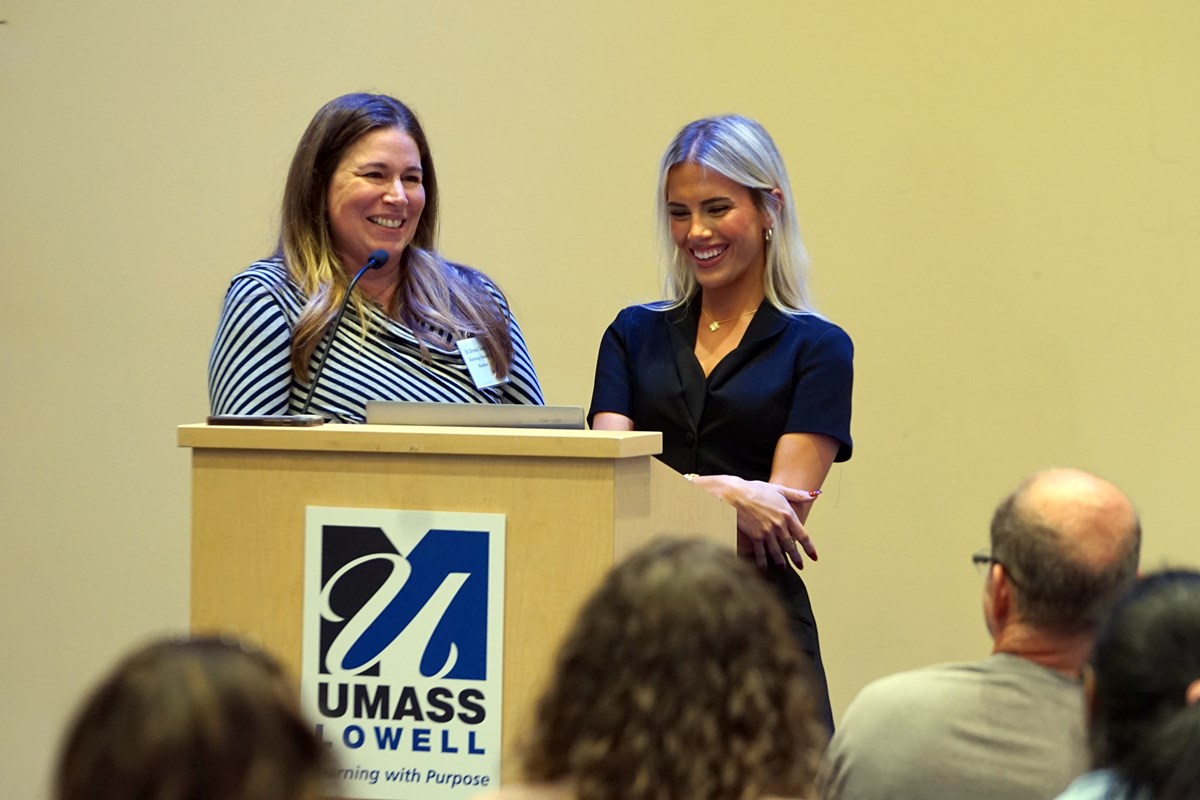 Two women smile while standing at a podium at the front of a room. There are several audience members seated before them.