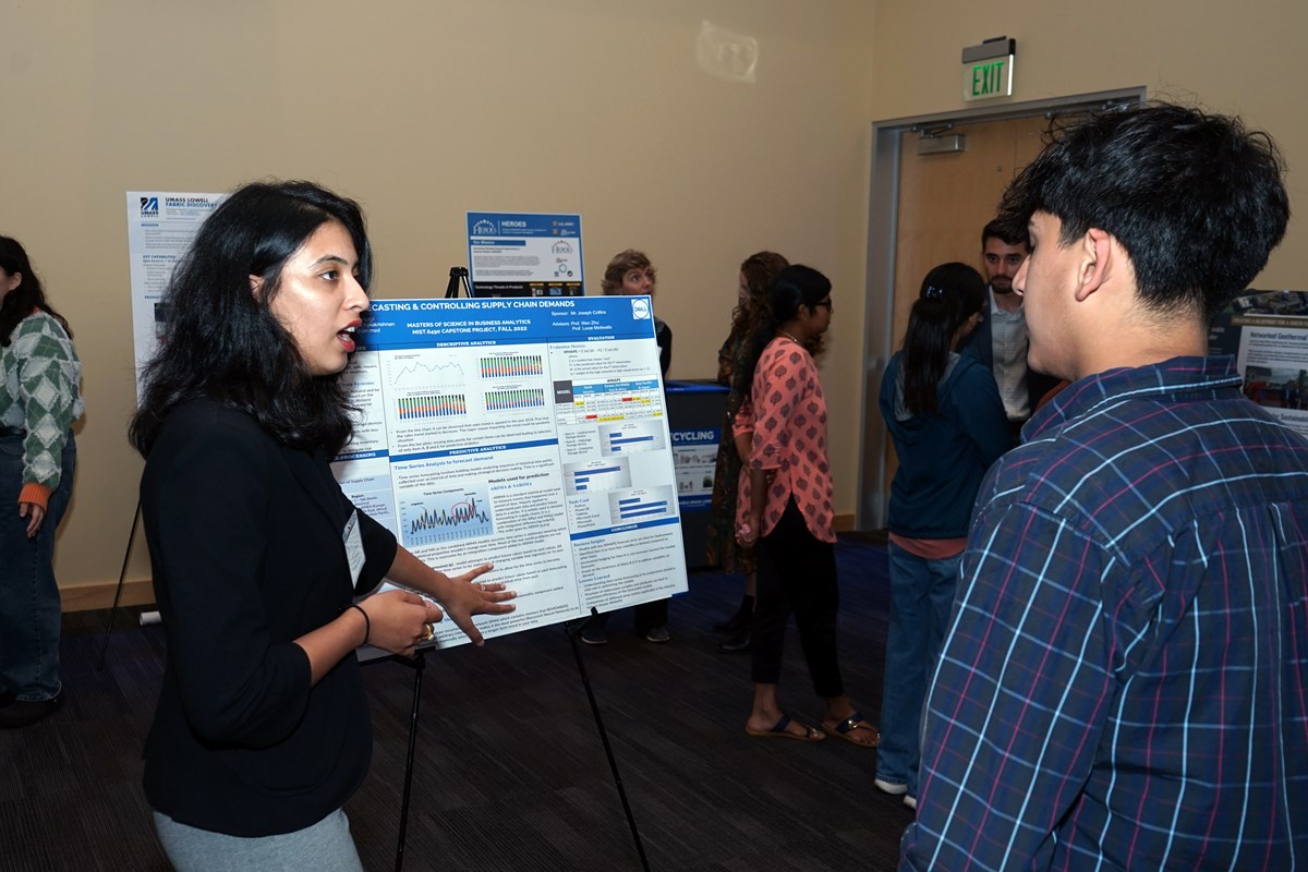 A young woman talks to a person while gesturing to a research poster mounted on an easel.