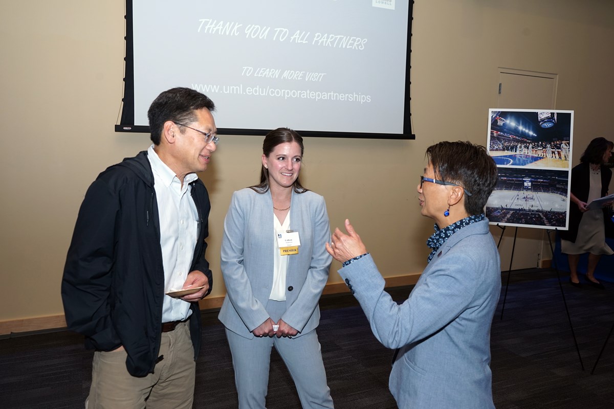 Three people talk to one another while standing in a room in front of a projection screen and a poster on an easel.