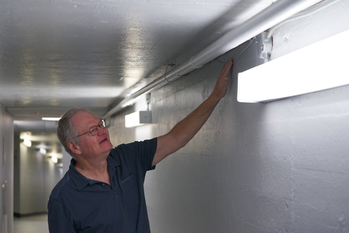 A man in glasses touches a wire that is running along the top of a wall in a tunnel.