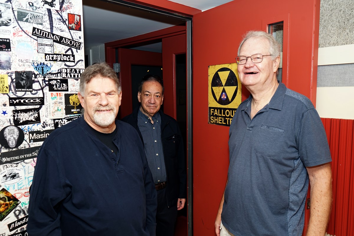 Three men pose for a photo in the doorway of a college radio station. There is a fallout shelter sign on the door.